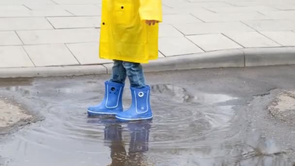 Bambino divertente con un impermeabile giallo e stivali di gomma che saltano in una pozzanghera, da vicino — Video Stock