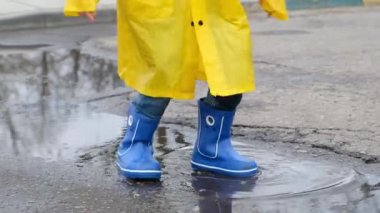 Sarı yağmurluk ve lastik çizme giyen komik çocuk su birikintisine atlıyor. Yaklaş.