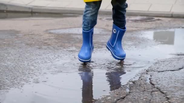 Grappig kind in een gele regenjas en rubberen laarzen springen in een plas, close-up, slow motion — Stockvideo
