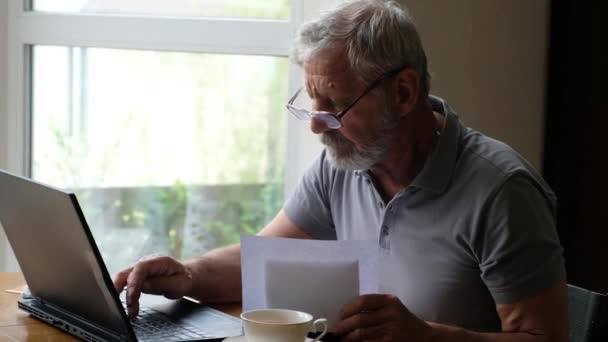 Senoir starzec w okularach siedzi przy stole i patrzy na laptopa, wpisując informacje z papieru — Wideo stockowe