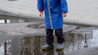 Mavi yağmurluklu ve lastik çizmeli komik çocuk su birikintisinde zıplıyor.