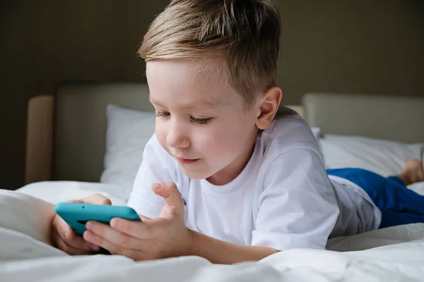 Lindo niño pequeño jugando juego móvil, acostado en una cama y sosteniendo el teléfono inteligente — Foto de Stock