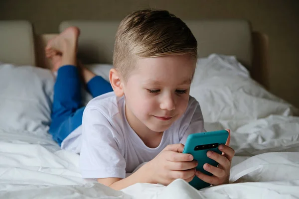 Lindo niño pequeño jugando juego móvil, acostado en una cama y sosteniendo el teléfono inteligente — Foto de Stock