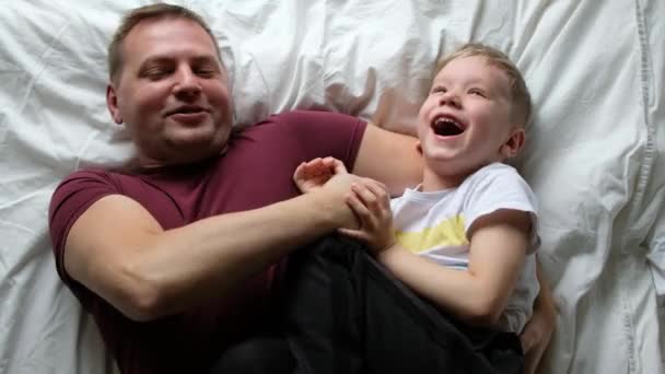 Lykkelig far som har det gøy i en seng med sønnen sin, ler. Sett ovenfra – stockvideo
