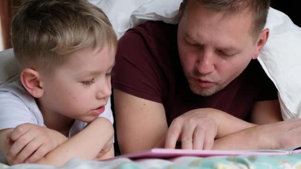 Far pappa och hans son ligger på en säng och läser en bok. Familjebegreppet — Stockvideo