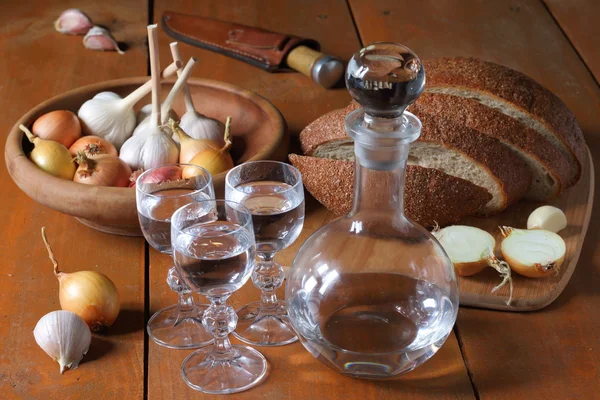 Jarra y tres vasos de vodka de pie sobre una mesa de madera junto al pan y las verduras . — Foto de Stock