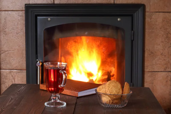 A glass of tea, biscuits and book are on the table in front of the fireplace.