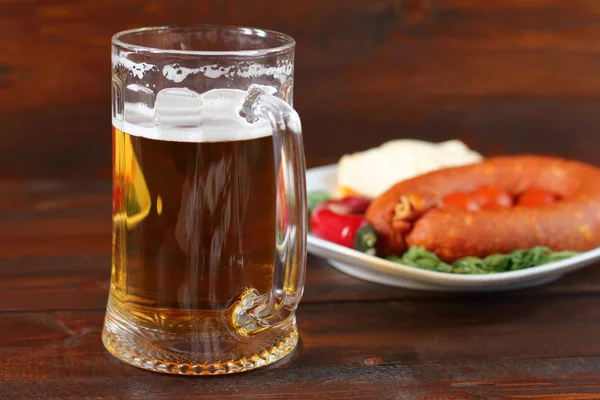 Кружка светлого пива и тарелка с копченой колбасой, сыр и овощи на темном деревянном фоне . — стоковое фото