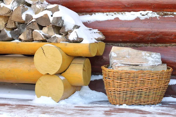 A wicker basket of firewood at the pile of birch firewood on the terrace of a country house in winter.