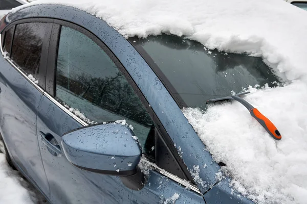 Nástroj na škrabání ledu pro čištění automobilů je na kapotě auta. Sníh odstraňuje z auta po sněhové bouři. Částečně vyčištěné čelní sklo. — Stock fotografie