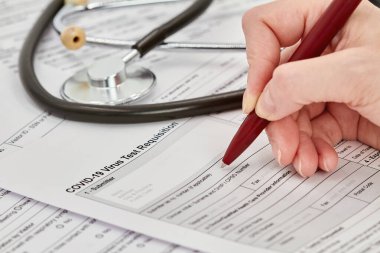 Doktor bir virüs testi talep formu doldurmaya hazırlanıyor. Arka planda steteskop var. Yakın çekim..