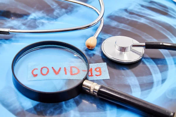 Stetoskop ve akciğer röntgeninin arkaplanındaki büyüteçle COVID-19 yazılıydı. Yakın çekim.
