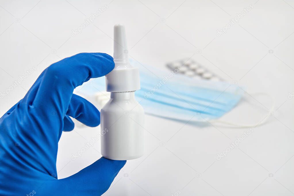A hand in blue protective glove holds a jar with nasal spray. Medical protective face mask, pills on the light background. Viral disease prevention concept.