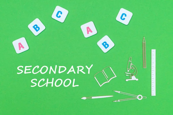 From above wooden minitures school supplies and ABC letters on green background with text secondary school — Stock Photo, Image