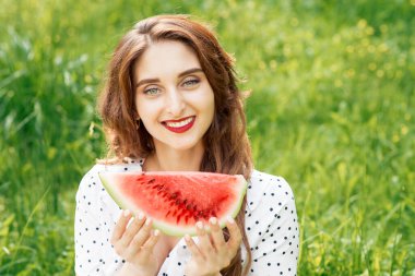 Güzel, gülümseyen bir kadın doğanın arka planında bir parça karpuz tutuyor. Genç, gülümseyen bir kadın elinde yeşil çimlerde karpuz parçası tutuyor..
