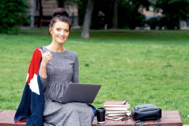 Kafkasyalı öğrenci dizüstü bilgisayar kullanıyor ve başparmağını parkta gösteriyor. Öğrenci kız dizüstü bilgisayarda çalışıyor ve başparmağını parktaki bankta gösteriyor..