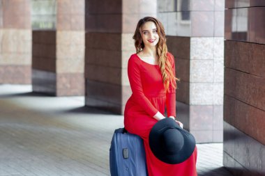 Siyah şapkalı güzel bir kadın bavula oturmuş kameraya bakıyor. Kırmızı uzun elbiseli genç gezgin kız bavulun üzerinde oturuyor..