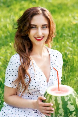 Beyaz elbiseli, gülümseyen bir kızın portresi yeşil çimenlerin arkasında kokteyl pipetli bir karpuz tutuyor..