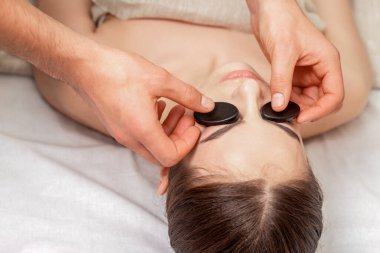 Genç bir kadın, kaplıcada mineral ve sıcak taşlarla yüz masajı yaptırıyor..