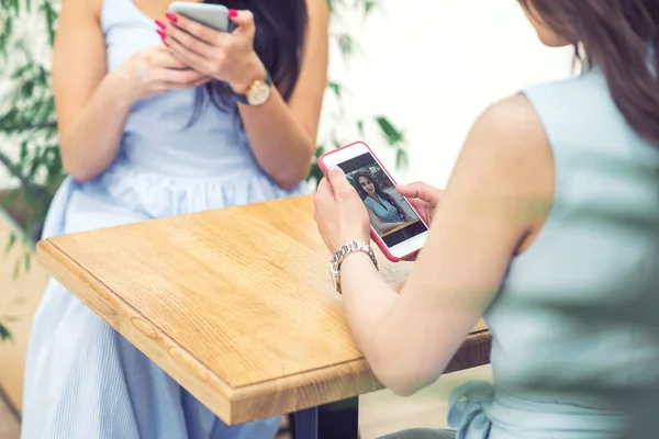 Two young women are looking at smart phone at cafe outdoors. Women sits in the street cafe with smartphone. Girl looks at herself on the smartphone.