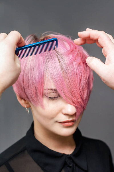 Hands of hairdresser are combing short pink hair of woman on gray background close up.