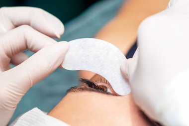 Kozmetik uzmanının elleri, kirpik uzatma işleminden önce genç bir kadının gözünün altındaki şeridi yapıştırır..