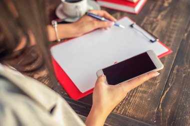 Bir kadının elleri deftere yazı yazıyor ve kafedeki ahşap masada akıllı telefon kullanıyor. Kadının elindeki siyah dokunmatik ekran.