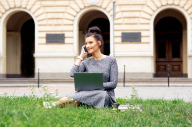 Öğrenci kadın üniversite binasının yakınındaki çimlerin üzerinde akıllı telefonla konuşuyor. Kız üniversitenin yanındaki yeşil çimlerde oturuyor..
