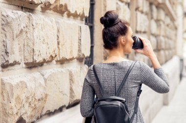 Taş duvarın yanında yürürken kağıt bardakta kahve içen bir kızın arkası. Genç bir kadın eski duvarın yanında yürüyor ve dışarıda kahve içiyor..