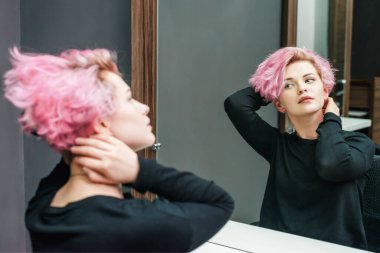 Saçlarını boyattıktan sonra aynaya bakan ve güzellik salonunda röfle yapan genç bir kadının güzel saç stili. Kısa pembe saç modelini kapat.