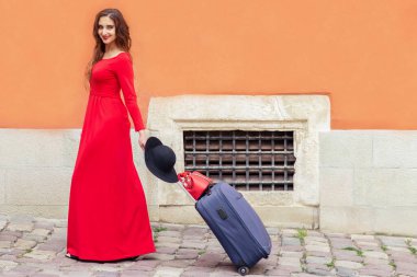 Güzel turist kadın şehir caddesinde bavulunu çekiyor. Uzun kırmızı elbiseli, bavullu, caddede yürüyen yetişkin bir kız..
