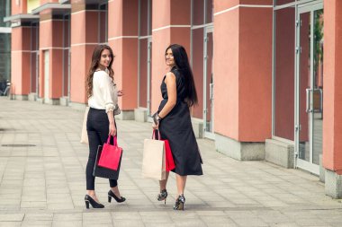 İki güzel kız alışveriş çantalarının arkasına bakarak şehir caddesinde yürüyorlar. Alışveriş yapan iki genç kadın. Boşluğu kopyala.