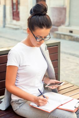 Güzel genç bir kadın sokakta bankta otururken defterine notlar alıyor. Yaşam Tarzı konsepti
