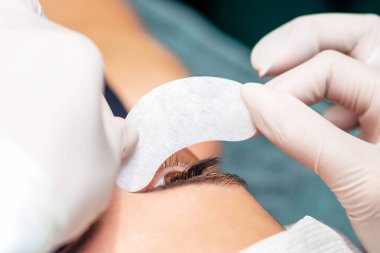 Kozmetik uzmanının yakın elleri, kirpik uzatma prosedüründen önce genç bir kadının gözünün altına şeridi koyar..