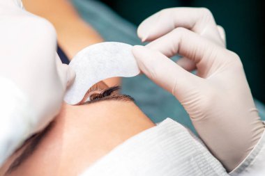 Kozmetik uzmanının yakın elleri, kirpik uzatma prosedüründen önce genç bir kadının gözünün altına şeridi koyar..