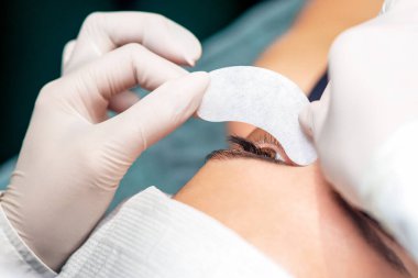 Kozmetik uzmanının yakın elleri, kirpik uzatma prosedüründen önce genç bir kadının gözünün altına şeridi koyar..