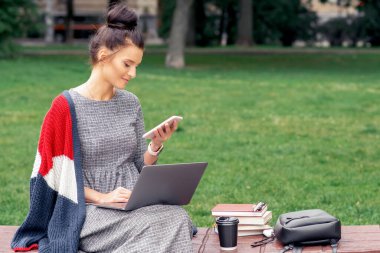 Öğrenci kız parktaki ahşap bir bankta dizüstü bilgisayarla akıllı telefona bakıyor. Bayan öğrenci dışarıda cep telefonu kullanıyor..