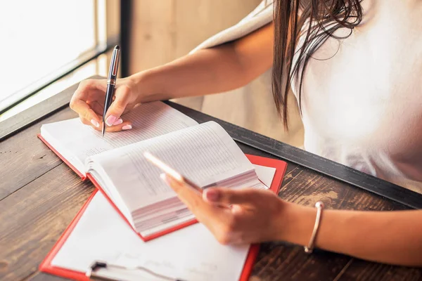 Genç iş kadını kafeteryada akıllı telefon tutarken deftere planlar yazıyor. İş konsepti