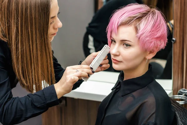 Hairdresser checking short pink hairstyle of young woman in hair salon.