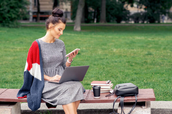 Student adult girl is looking on the smartphone at the park. Female student is looking on cell phone with laptop while sits on the bench at the park.