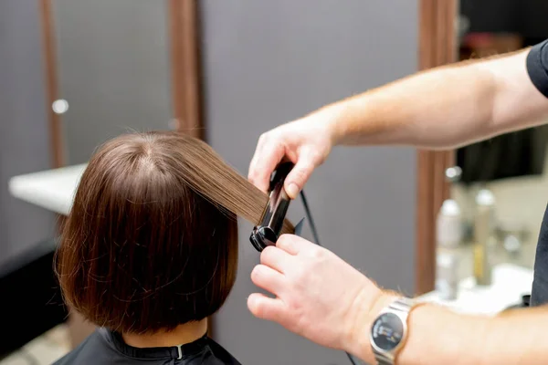 Hairdresser straightens hair of woman with hair straightener tool in hair salon.