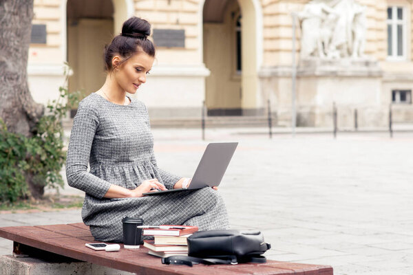 A young pretty woman works on laptop computer near building of university. Female student is working on laptop computer outside university building.