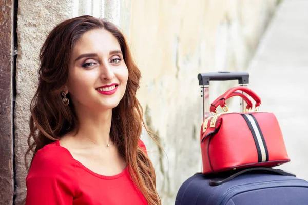 Portrait of beautiful smiling woman with suitcase in red dress over old wall.