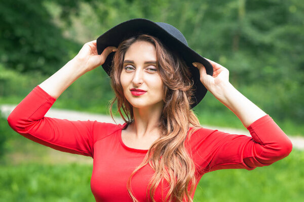 Young woman is walkinig in black hat at the summer park. Brunette girl in black hat and red sweater on green nature.