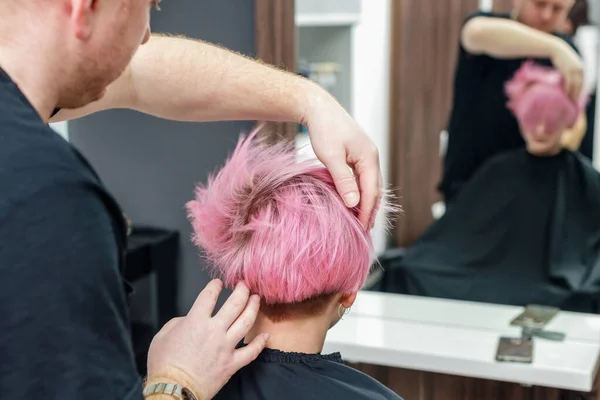 Hairdresser is checking short pink hairstyle of young woman in the hair salon.