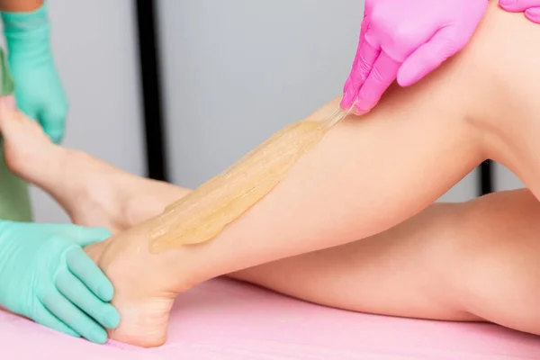 Beautician's hand is applying liquid sugar wax on leg during of sugaring epilation process.