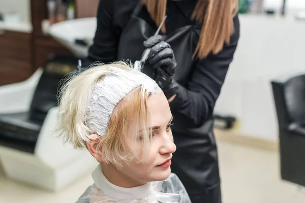 Hairdresser in black gloves applies white dye on woman\'s hair in beauty salon.