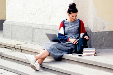 Basamaklarda dizüstü bilgisayar ve sırt çantasıyla oturan çekici bayan öğrenci. Öğrenci kız merdivenlerde sırt çantasında bir şey arıyor..