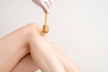 Wax ax flows down the honey stick on leg during hand holding stick on white background. Concept of depilation and epilation. clipart