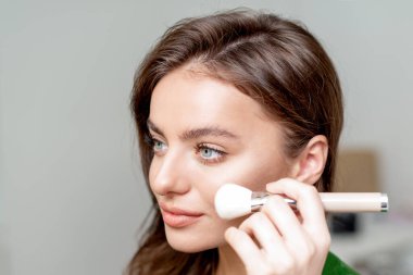 Makyaj fırçası kullanan bir kadının yakın plan portresi yüzüne kuru kozmetik ton temeli uyguluyor..